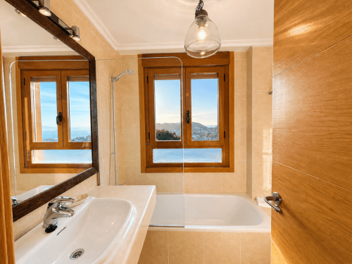 Baño del apartamento vacacional en Baiona con vistas al mar, puertas de madera, lavabo blanco, bañera y vista del Océano Atlántico