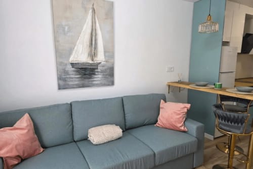 Acogedora sala de estar en Smart Rent Fuerteventura con sofá turquesa, cojines rosas, cuadro de velero y área de cocina
