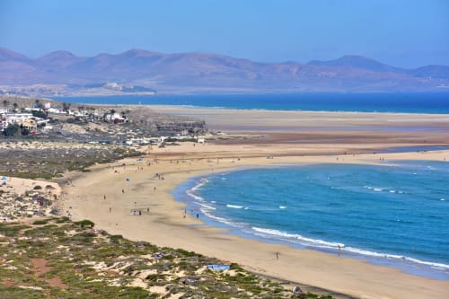 Vista aérea espectacular de la playa de Fuerteventura con aguas turquesas, arena dorada y montañas bajo cielo azul despejado