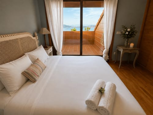 Dormitorio de lujo con balcón vista al mar en Baiona, ropa de cama blanca, suelos de madera y vistas al Océano Atlántico a través de puertas de cristal