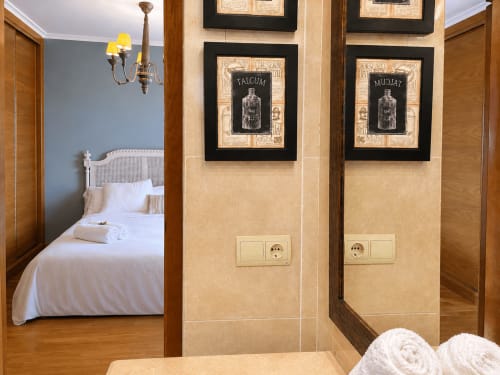 Entrada del baño del apartamento Baiona con paredes beige, cuadros de botellas vintage, puertas de madera y toallas enrolladas.