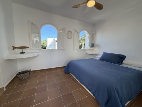 Dormitorio luminoso en Villa Alegria de Cala d'Or con cama azul, ventanas arqueadas, pisos de terracota y ventilador de techo