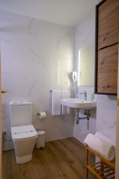 Baño moderno del Hotel Boutique Alcazaba con paredes de mármol, espejo iluminado, lavabo blanco, inodoro y toallero de madera