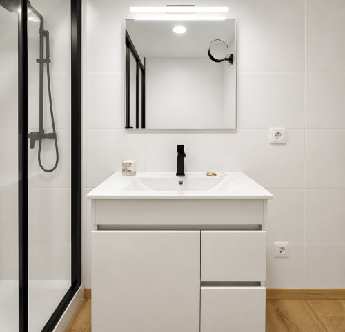 Baño moderno en apartamento de Baiona con vanidad blanca, accesorios negros, espejo con marco y ducha abierta con marco negro