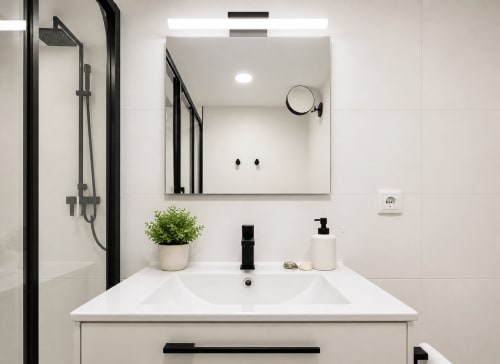 Moderno lavabo de baño con espejo en el apartamento Holibai en Baiona, España. Griferías negras, encimera blanca, planta decorativa.