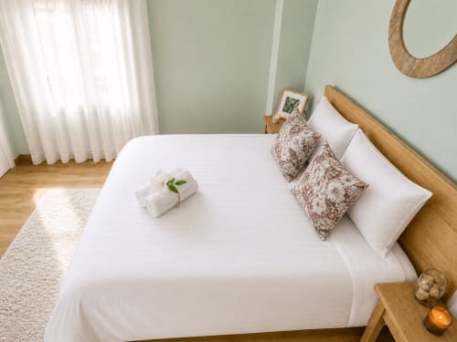 Dormitorio luminoso en apartamento de Baiona con sábanas blancas, cama de madera, almohadas estampadas y vistas a la ciudad por ventana con cortinas