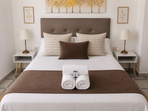 Apartamento Holibai en Baiona con cama tendida, cabecero marrón, almohadas blancas, toallas enrolladas y lámparas de mesita