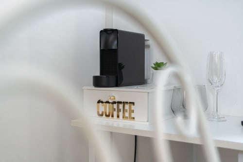 Macchinetta del caffè nera su ripiano bianco con scritta 'COFFEE', bicchieri e pianta grassa nella spa dell'Elite Apartment