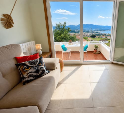 Luminosa sala de estar con sofá beige, cojín de búho y puertas de cristal que abren a terraza con vistas a Playa América y montañas