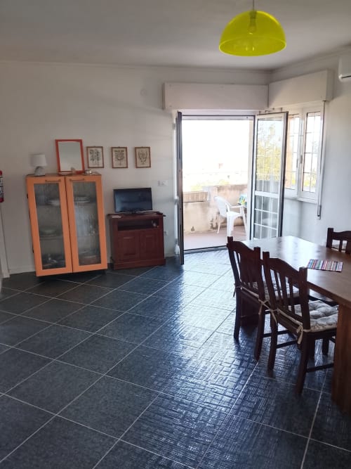 Spazioso soggiorno con zona pranzo, mobile arancione, pavimento in piastrelle scure, lampada a sospensione gialla, e porte in vetro che si aprono su