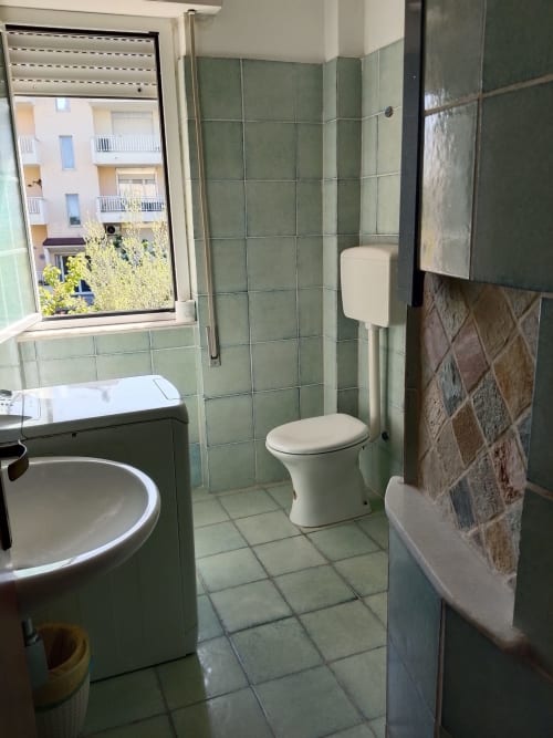 Bagno luminoso dell'appartamento Tales of Abruzzo Fossacesia con piastrelle verdi, toilette, lavandino e finestra con vista sulla strada abruzzese