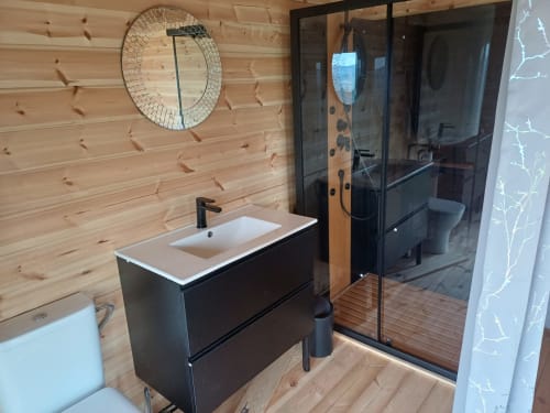 Modern bathroom at La Maison de Dun with wooden walls, black sink vanity, glass shower enclosure, and round mirror