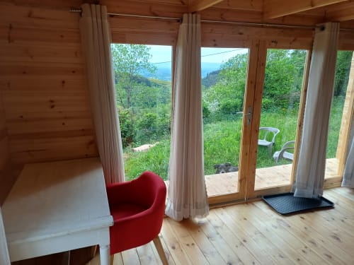 Wooden cabin interior with red chair, open glass doors overlooking green mountains at La Maison de Dun, Saint Racho