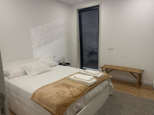 Quarto luminoso na MiraReal GuestHouse em Macieira da Lixa com cama branca, banco de madeira, venezianas ensolaradas e piso de madeira