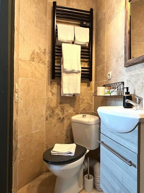 Salle de bain moderne du gîte Maisons Delphine avec porte-serviettes chauffant, lavabo blanc, toilettes et carrelage en travertin