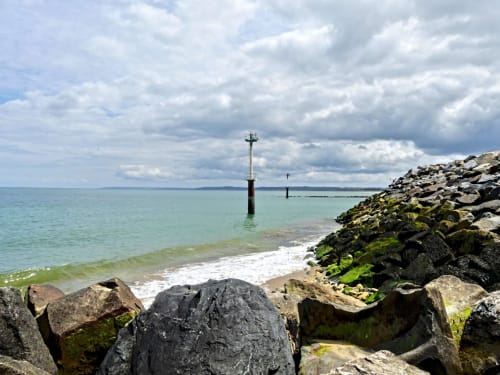 Rocky coastline at Lion sur Mer with calm turquoise waters, offshore navigation markers, and cloudy sky