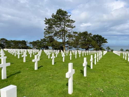 American cemetery with white crosses on green lawn, trees and sea visible in background, Lion sur Mer