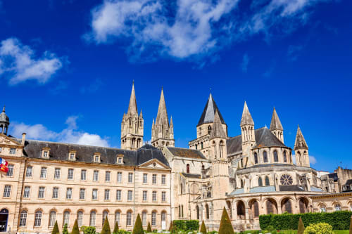 Ornate cathedral with multiple spires and stone architecture beside cream-colored building in Lion sur Mer, France