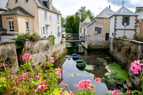 Picturesque canal in Lion sur Mer with moored boats, stone buildings, pink flowers, and weeping willow trees