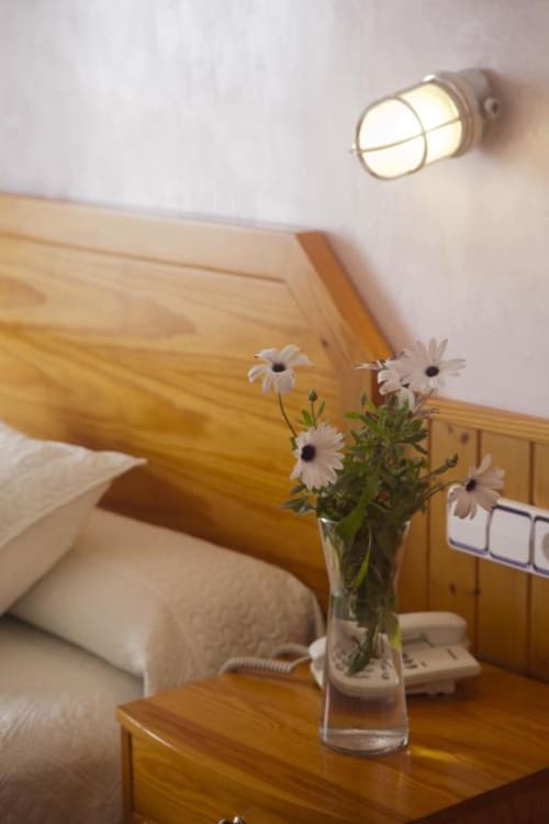 Acogedora habitación en Hostal Mar de Frente con cama de madera, margaritas blancas en jarrón de cristal en la mesita de noche y lámpara de pared