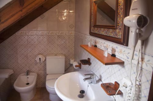 Baño rústico en buhardilla del Hostal Mar de Frente con azulejos blancos, vigas de madera, lavabo, inodoro y espejo decorativo.