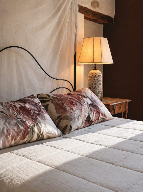 Chambre cosy du gîte Viento de Ladera à Ségovie avec coussins floraux, draps blancs, lampe de table et plafond en bois rustique