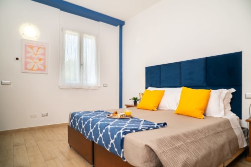 Camera da letto moderna con testata blu, cuscini gialli e vista sul giardino dell'appartamento Match Home a Somma Lombardo