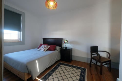 Quarto luminoso no Santiago de Caminha Boutique Hostel com cama, criado-mudo preto, luminária pendente, janela e piso de madeira.