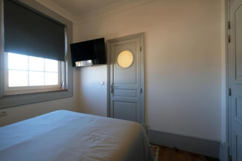 Quarto moderno na Santiago de Caminha Boutique Hostel com cama, TV na parede, janela com persiana e porta azul claro