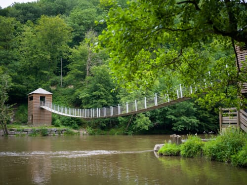 Suspended footbridge over river at Aux 4 Chalets Corbion, Bouillon, surrounded by lush green forests