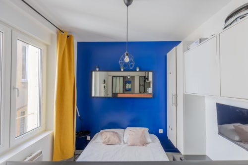 Studio moderne à Metz avec mur accentué bleu, lit, téléviseur et rideaux jaunes près de la fenêtre