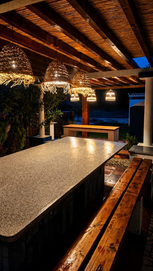 Cozinha externa com bar na Herdade da Cova da Onça, luminárias de vime iluminadas, pérgola de madeira e vista para o oceano ao entardecer