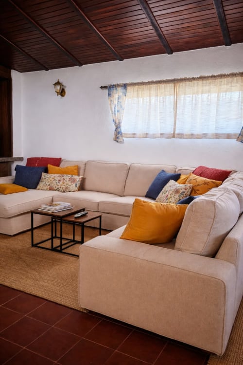 Aconchegante sala de estar da Herdade da Cova da Onça com sofá secional bege, almofadas coloridas e teto em madeira