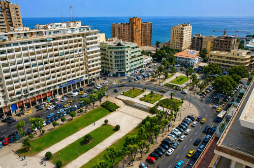 Aerial view of DakarByDays hotel area showing beachfront buildings, green plaza, parked cars, and Atlantic Ocean in Dakar