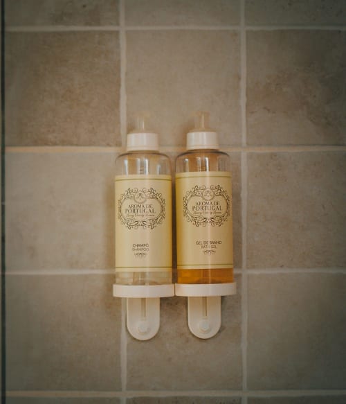 Frascos de champô e gel de banho Aroma de Portugal em dispensador branco na parede do banheiro em azulejos da Herdade da Cova da Onça