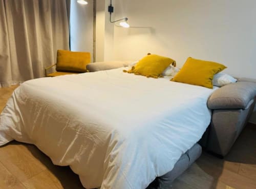 Dormitorio moderno en apartamento Gestyp-AT con cama blanca, almohadas amarillas, cabecera gris, piso de madera y luz colgante