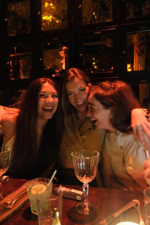Three women laughing together at a dinner table with drinks at Riad 191 in Marrakech, warm ambient lighting and windows