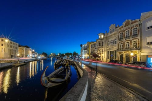 Vista noturna do canal de Aveiro com barcos tradicionais, edifícios históricos iluminados e rua de paralelepípedos na margem
