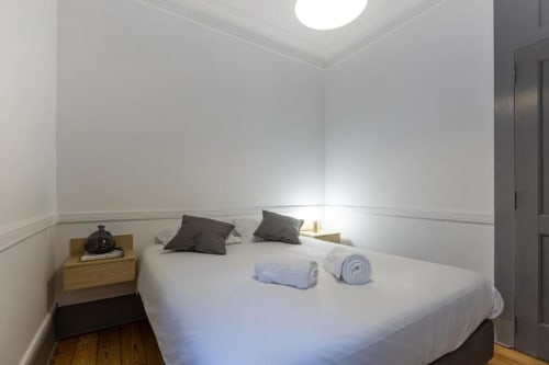 Quarto moderno no Aveiro com Paixão Living com cama branca, almofadas escuras, toalhas enroladas, criado em madeira e teto iluminado