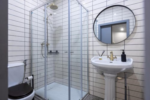 Banheiro moderno com azulejos brancos subway, pia coluna, espelho redondo e chuveiro com box em apartamento português