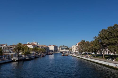Canal de Aveiro com edifícios históricos, barcos e passeio à beira-rio sob céu azul claro
