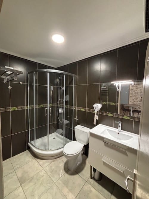 Salle de bains moderne de l'hôtel Le Clos Androne à Sisteron avec carrelage foncé, douche arrondie, vasque blanche et luminosité naturelle
