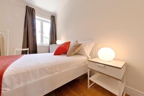 Quarto moderno com cama branca, almofada vermelha, piso de madeira e vista para a cidade no apartamento do Porto