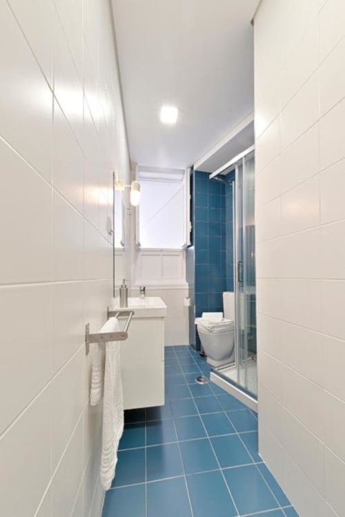 Banheiro moderno em apartamento no Porto com azulejos brancos nas paredes, piso em azulejos azuis, pia, vaso sanitário e box de vidro com vista para a cidade