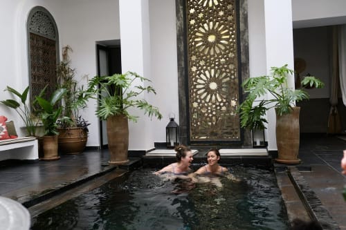 Two guests enjoying a dark water pool at Riad 191 in Marrakech, surrounded by lush plants and ornate Islamic architecture