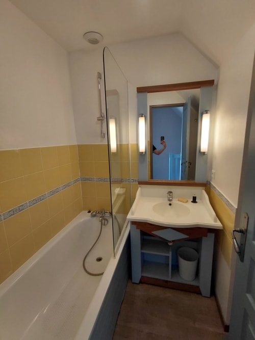 Salle de bains moderne aux murs carrelés jaunes, lavabo blanc, baignoire avec douche vitrée et miroir éclairé à Locmaria