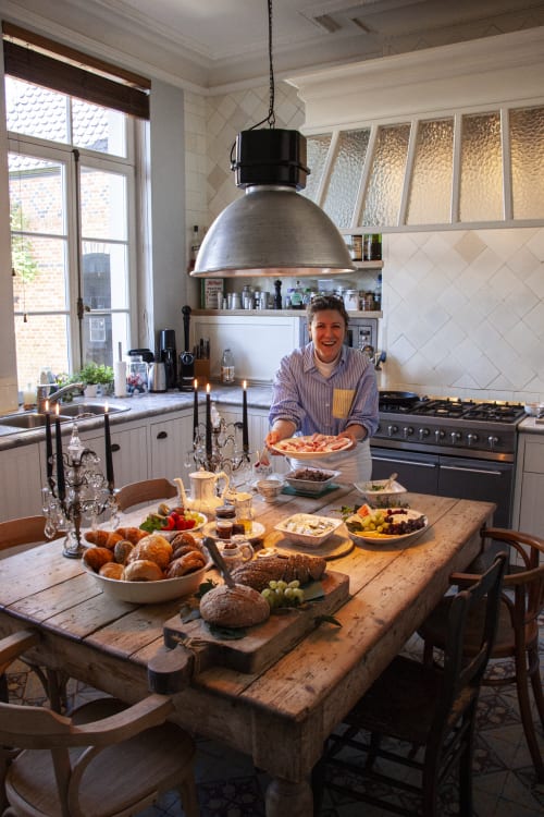 Host smiling in elegant kitchen with rustic wooden table displaying fresh pastries and food at Castanea Business & Suites,