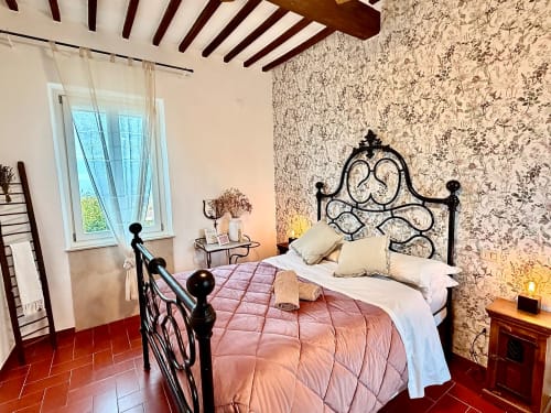 Elegant bedroom at Agriturismo CasaViVa with ornate black iron bed, pink bedding, floral wallpaper, wooden beams, and garden