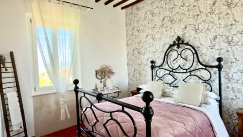 Camera da letto presso l'Agriturismo CasaViVa con letto in ferro battuto ornamentale, biancheria rosa, carta da parati floreale e vista sul giardino.