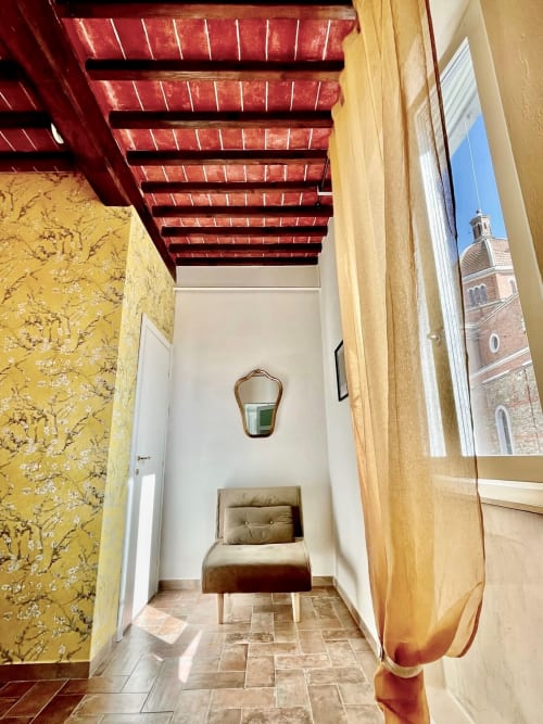 Corridoio stretto con pareti a motivi gialli, travi rosse a vista, poltrona beige e vista del campanile italiano dalla finestra